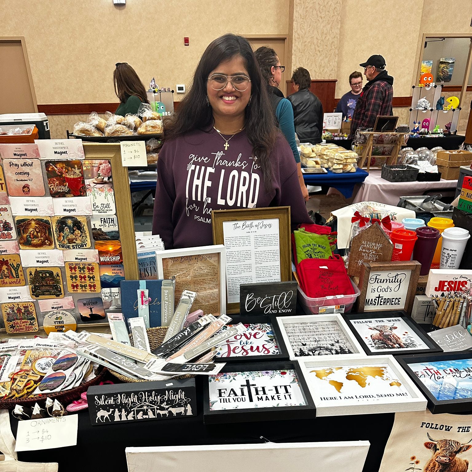 Woman standing behind a table with various items and a sign reading 'Bethel Masterpiece' at an event.