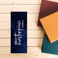 Navy blue bookmark with 'GOD'S MASTERPIECE' text on a wooden surface with books.