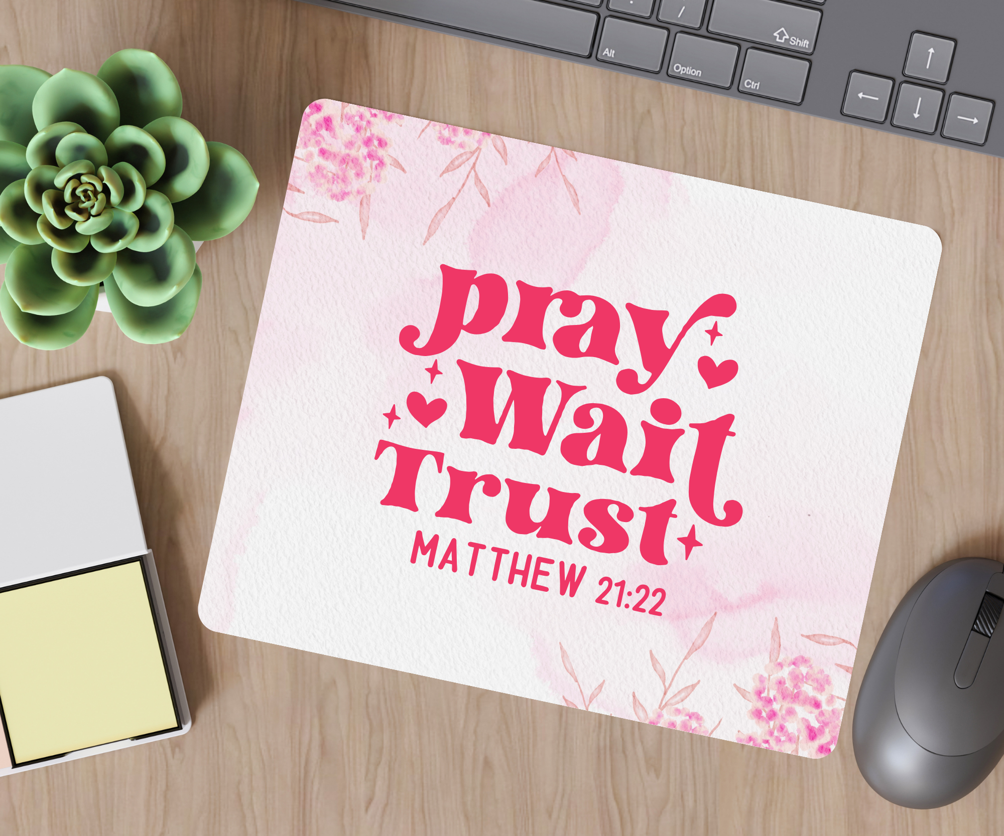 Desk setup with a mousepad featuring 'pray Wait Trust' text, keyboard, mouse, and plant.